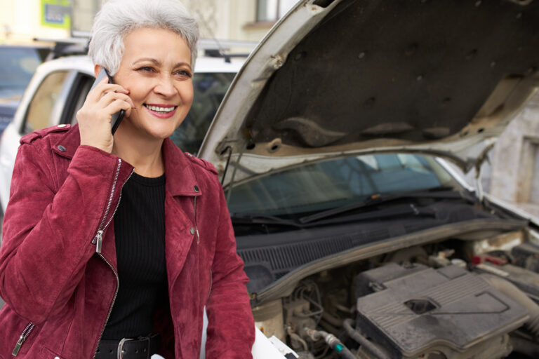 Cheerful stylish middle aged female having her white automobile being serviced during inspection, smiling broadly, making phone calls. Modern retired woman with mobile posing near car with open hood