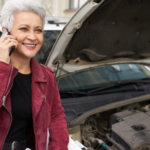 Cheerful stylish middle aged female having her white automobile being serviced during inspection, smiling broadly, making phone calls. Modern retired woman with mobile posing near car with open hood